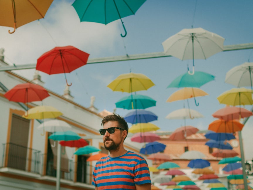 A man wearing sunglasses and a striped shirt standing under a canopy of colorful hanging umbrellas, with buildings and a clear sky in the background.